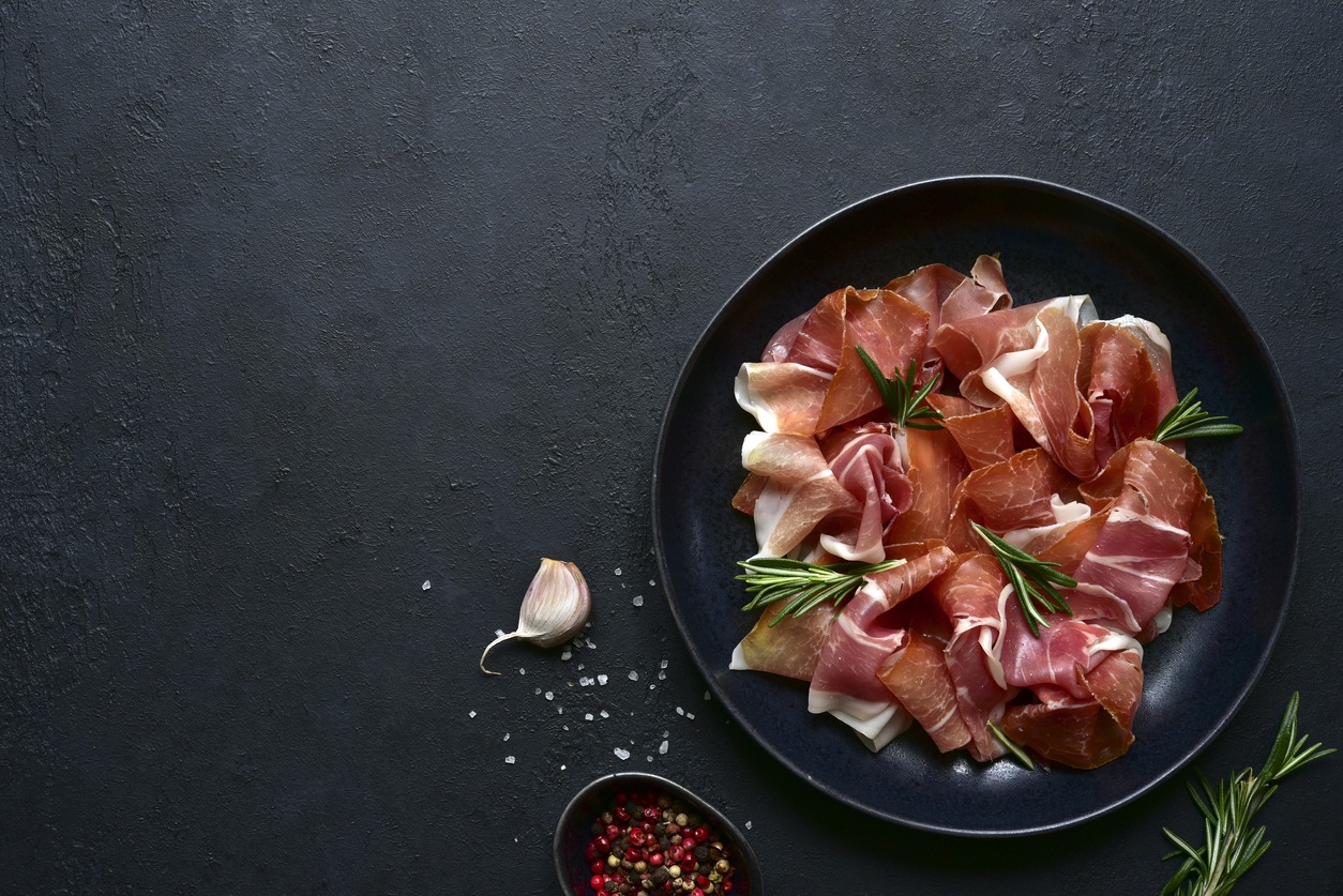Slices of prosciutto di parma or jamon serrano (iberico) on a black plate on a dark slate, stone or concrete background