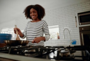 Low-angle view of young smiling woman mixing ingredients and vegetables in pan while preparing lunch