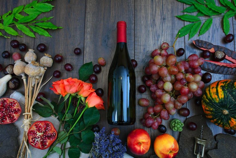 a wine bottle surrounded by grapes, roses, and several fruits on a brown wooden surface